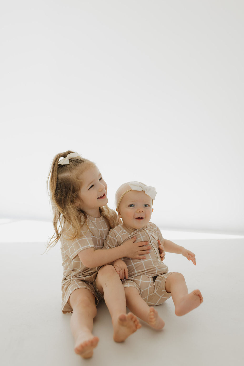 Two young children sit on a white surface in matching Bamboo Short Sets from forever french baby. The older child’s long hair is tied with a bow, while the younger wears a headband. Their smiles radiate happiness as they enjoy the comfy, breathable fabric perfect for sensitive skin.