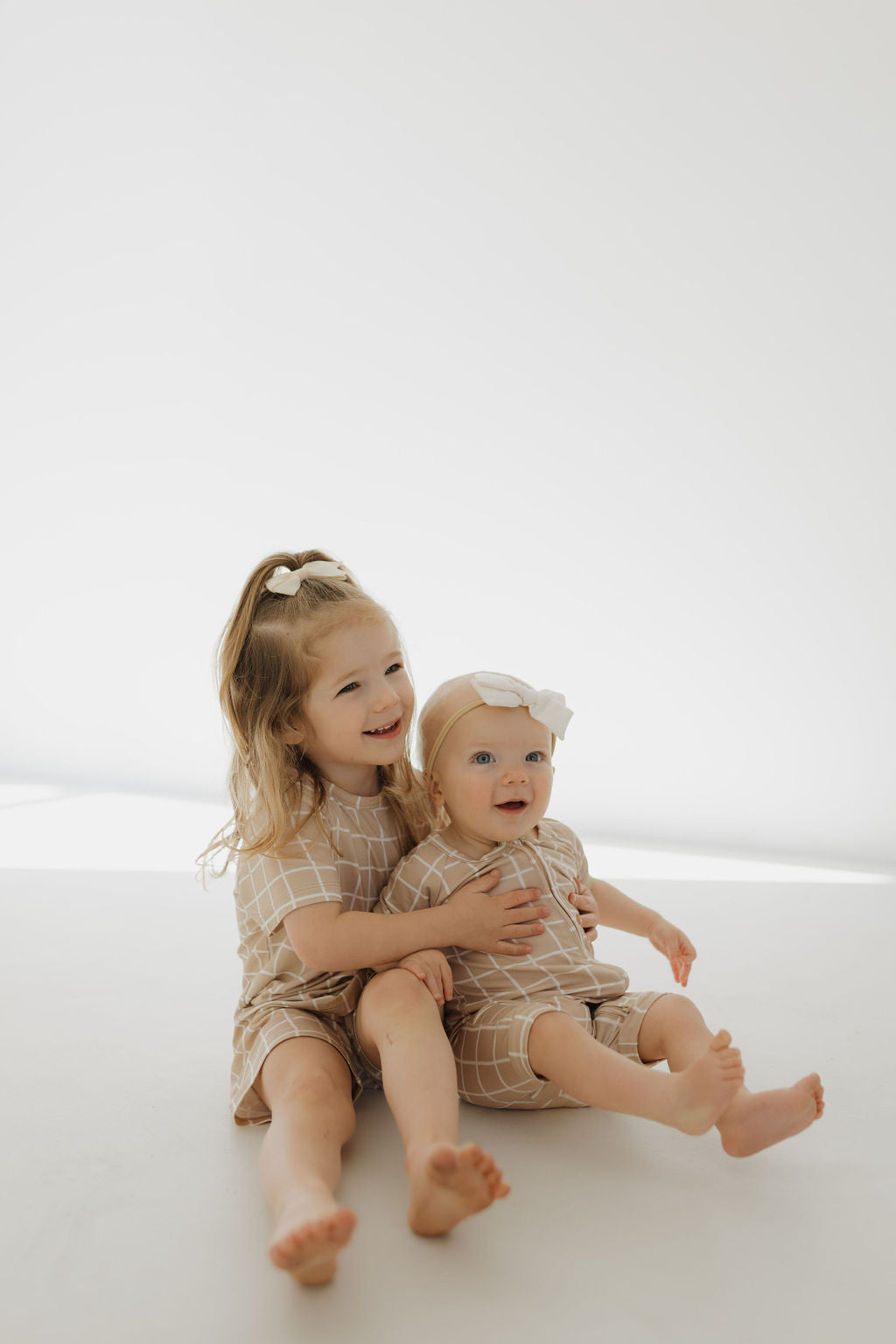 Two young children sit on a light floor in matching forever french baby Bamboo Short Sets | Grid. The older, with long hair and a bow, hugs the younger wearing a headband. Both are barefoot and smiling in their hypo-allergenic, breathable outfits in a softly lit room.