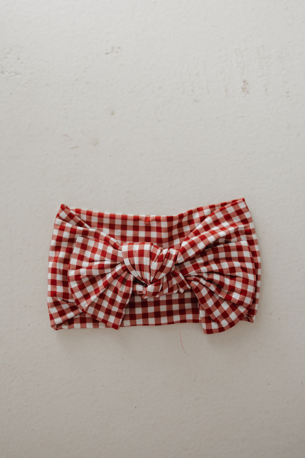 The Bamboo Head Wrap in Red Gingham from forever french baby features a large center bow and soft fabric, displayed on a light textured surface—an adjustable, stylish accessory for any occasion.
