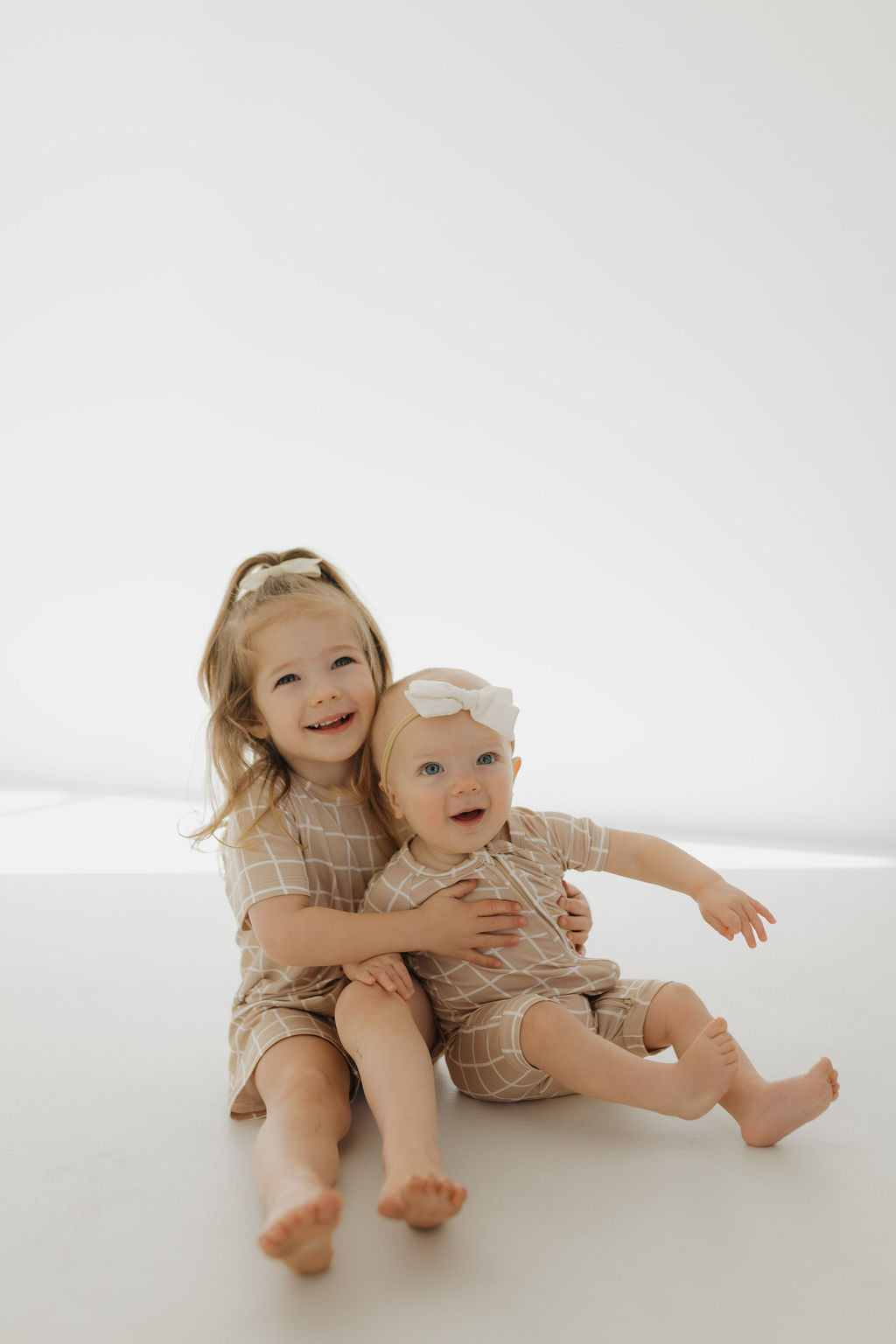 Two young children sit on the floor against a light background, dressed in matching Grid outfits from forever french baby’s Bamboo Zip Shortie collection. Both are smiling; the older child has long hair, and the younger one wears a bow headband.