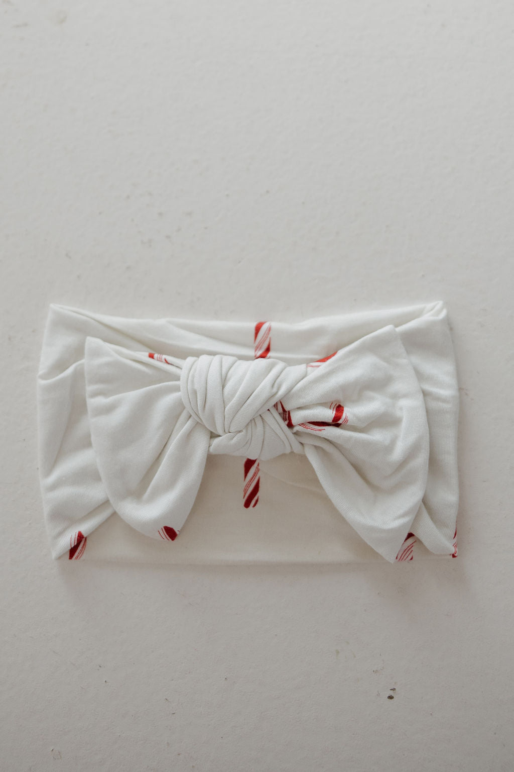 The Bamboo Head Wrap | Candy Cane by forever french baby is a white baby head wrap with red candy cane stripes and a large knotted bow, shown on a neutral background. A cute and popular accessory for little ones.