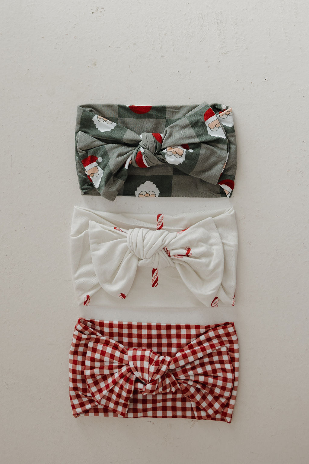 Three knotted fabric headbands from forever french baby are laid out vertically: the top is green with Santa print, the middle white with candy cane stripes, and the bottom is the Bamboo Head Wrap in Red Gingham, all on a light background.