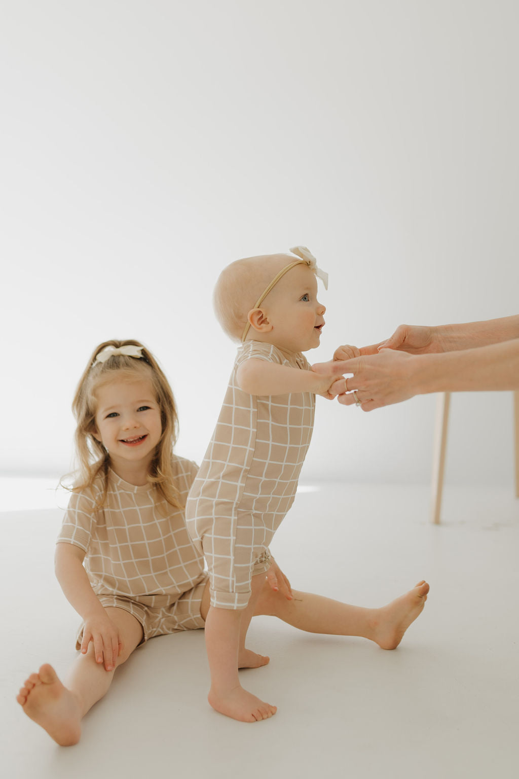 In a bright room, a toddler and baby wear matching Bamboo Short Sets with a Grid design from forever french baby. The toddler is seated and smiling while the baby stands, supported by adult hands, creating a light and hypo-allergenic scene.