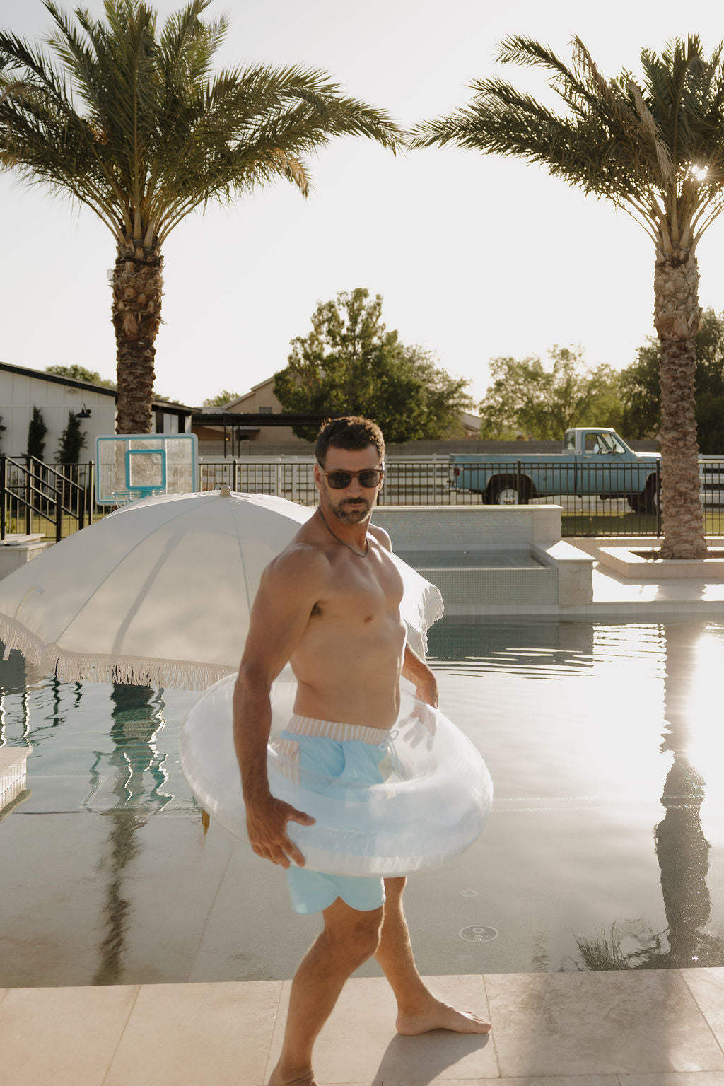 A man by the pool on a sunny day sports lolo webb 2 Men Board Short | Surf & Sand with sunglasses, a clear inflatable tube around his waist, palm trees, and a white umbrella in the background.