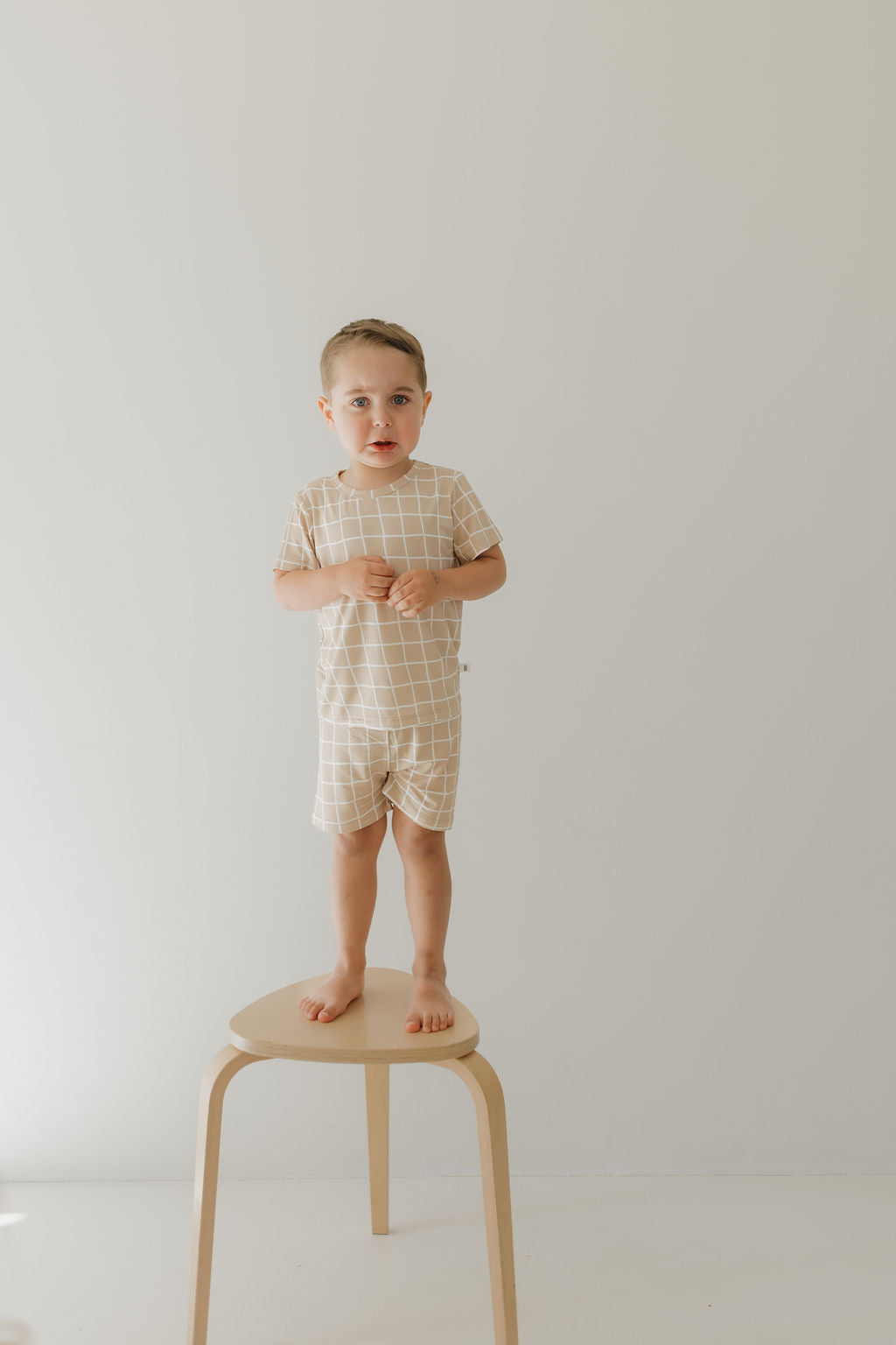 A young child stands barefoot on a wooden stool against a light background, wearing the Bamboo Short Set | Grid by forever french baby. The beige and white grid pattern provides breathable comfort. The child looks forward confidently.