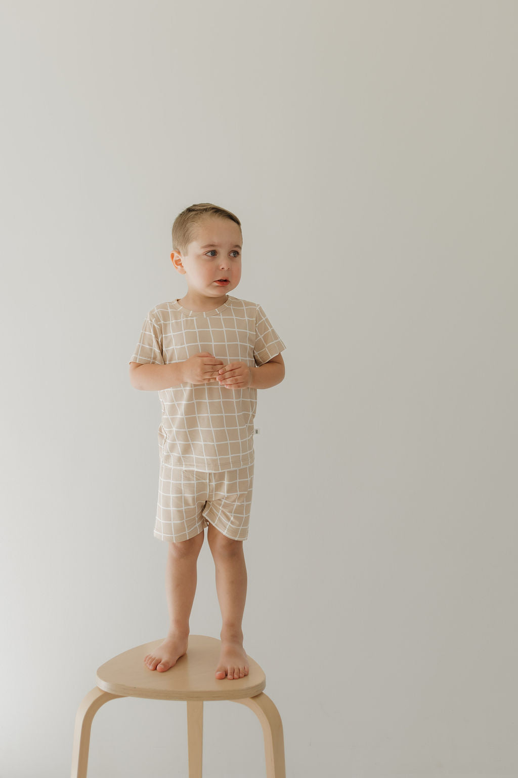 A young child stands barefoot on a wooden stool against a plain light background, wearing the breathable Bamboo Short Set | Grid by forever french baby in beige and white checks. This hypo-allergenic outfit complements the child's neutral expression while gazing to the side.