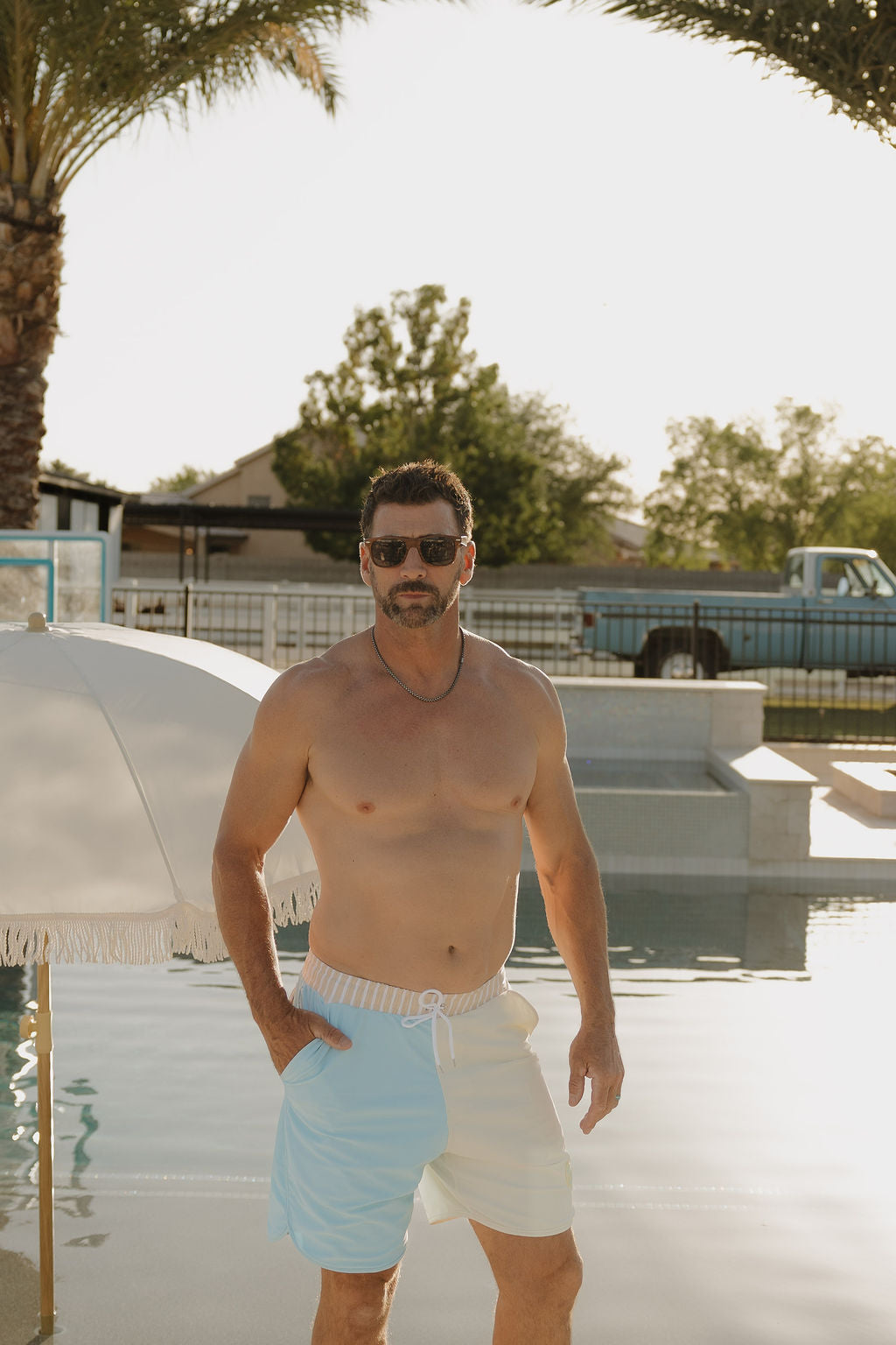 A man in lolo webb 2's limited edition Men Board Short | Surf & Sand stands poolside on a sunny day, with yellow and white stripes, palm trees, a white umbrella, and a vintage blue truck in the background.