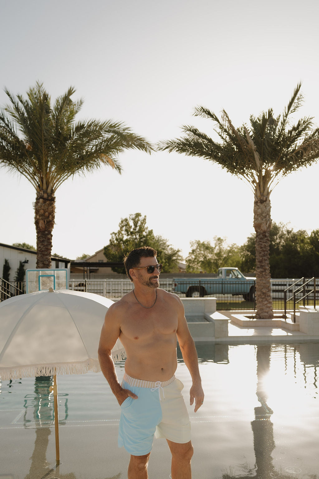 A man in lolo webb 2 Men Board Short | Surf & Sand stands poolside, wearing sunglasses, with palm trees and a white umbrella in the sunny background.
