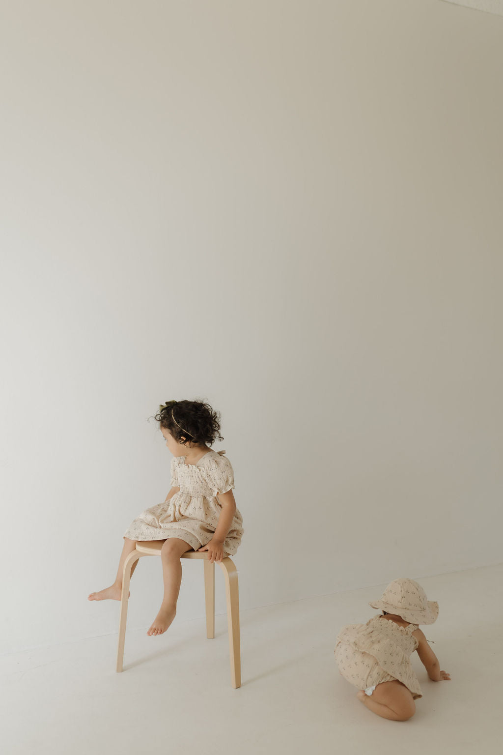 Two young children in forever french baby outfits; one wears the Toddler Muslin Dress | Flora, seated on a stool and looking left. The other, in a sun hat, crawls nearby. A minimal, softly lit backdrop captures sweet mom-and-me moments.