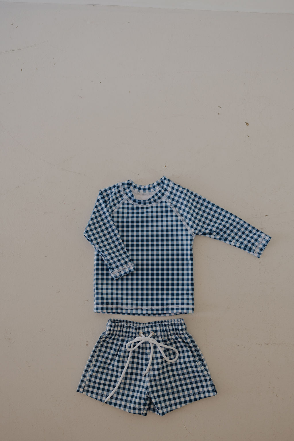 The forever french baby Child Board Short in Blue Gingham is a two-piece kids' summer set with a long-sleeve top and matching board shorts featuring a white drawstring, shown laid flat on a light background.