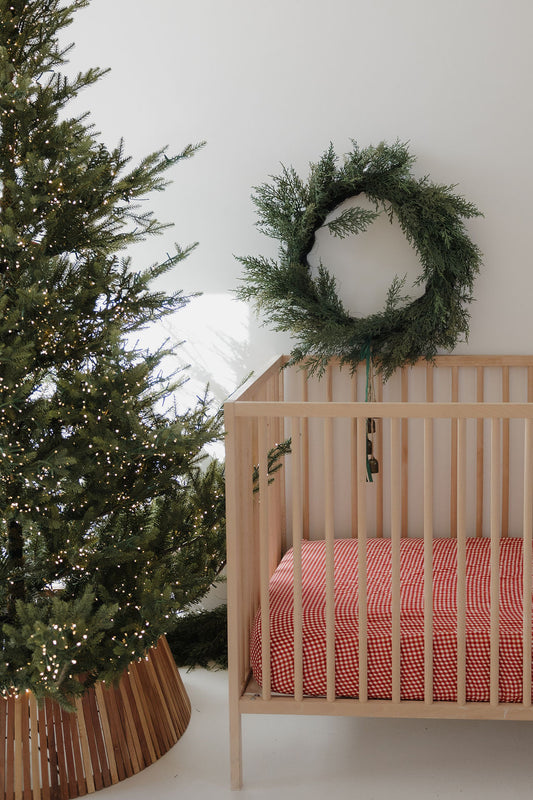 A forever french baby Muslin Crib Sheet in Red Gingham covers a wooden crib beside a Christmas tree and green wreath, adding charming baby bedding to a festive, minimalist holiday scene.
