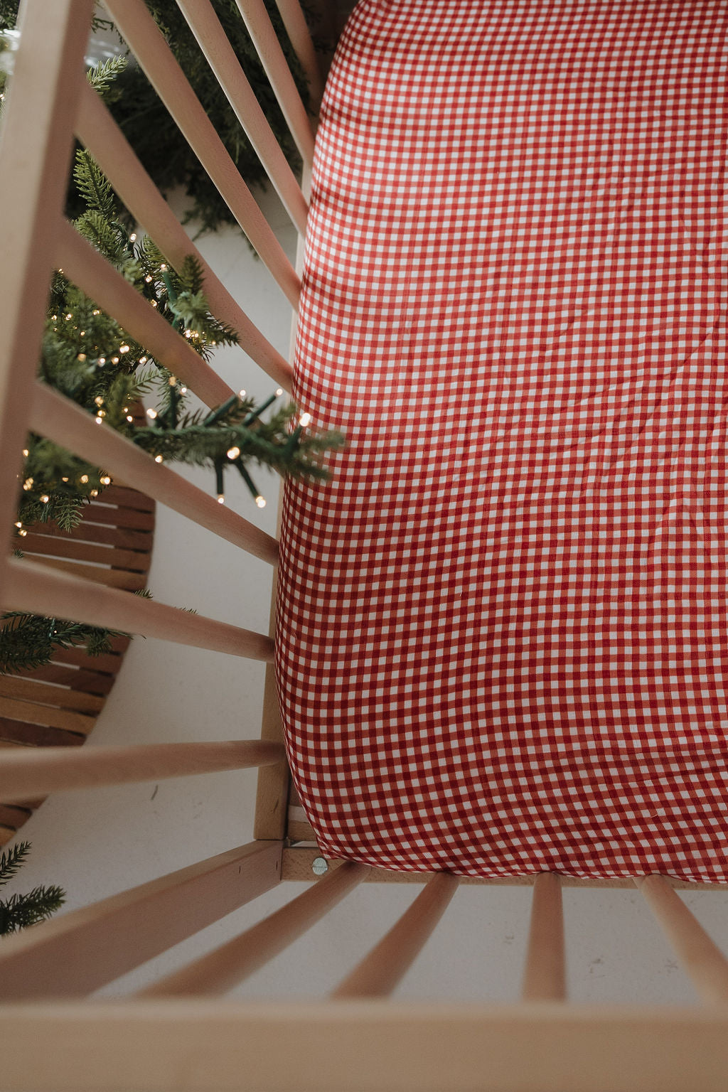 A top view of a wooden crib featuring the forever french baby Muslin Crib Sheet in Red Gingham, with green pine branches and string lights along the side for a cozy, inviting nursery look.