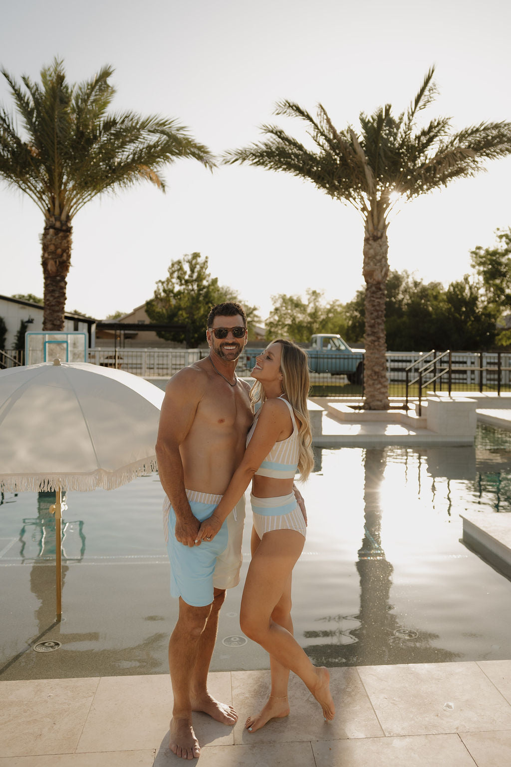 A smiling couple stands by the pool holding hands, wearing lolo webb 2's Women Two Piece Swimsuit | Surf & Sand. Palm trees and a white umbrella in the background create the perfect sunny vibe.
