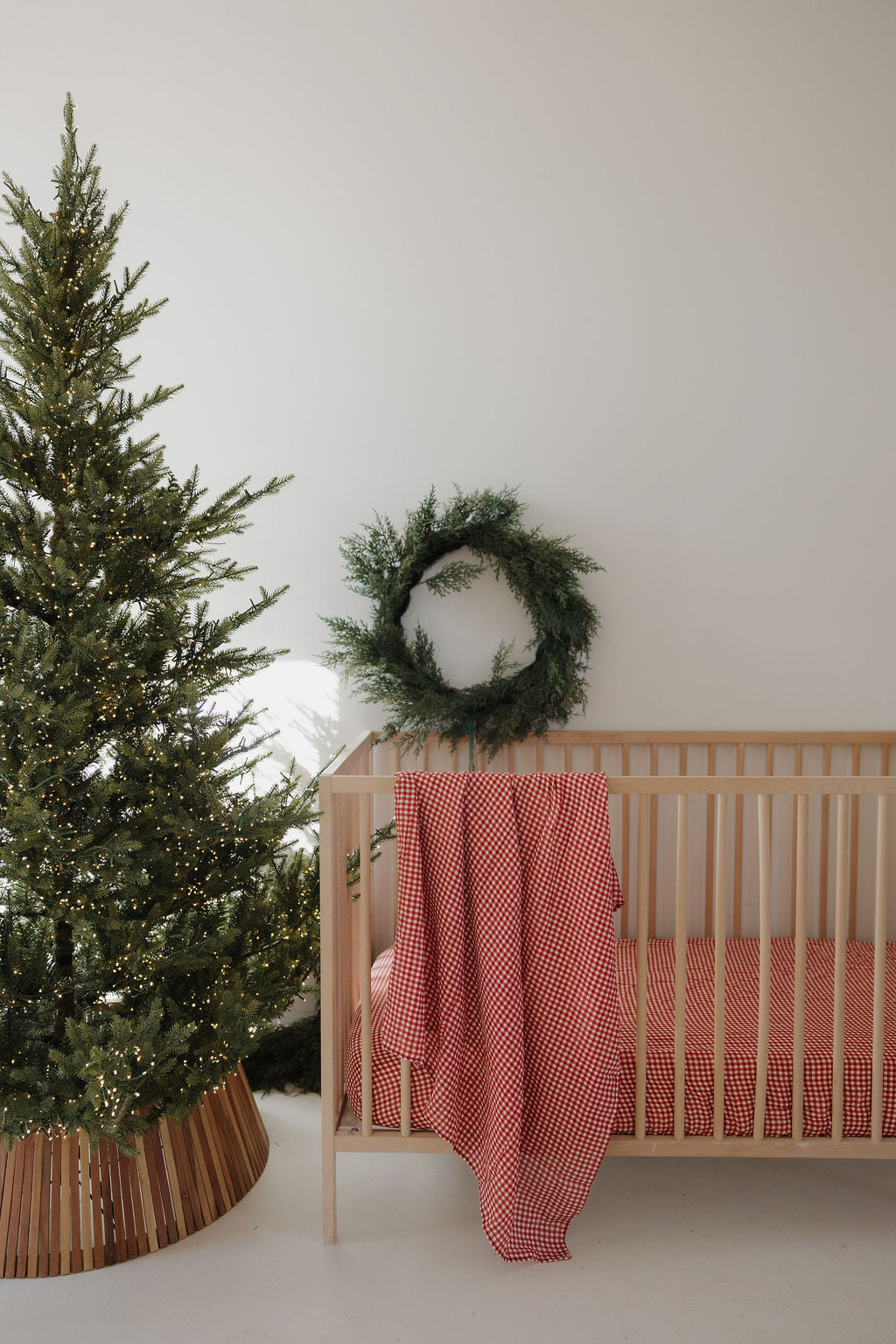 A simple nursery features a wooden crib with red bedding, a forever french baby Muslin Swaddle | Red Gingham, a red and white blanket draped on the crib, a decorated Christmas tree, and a green wreath on the white wall.