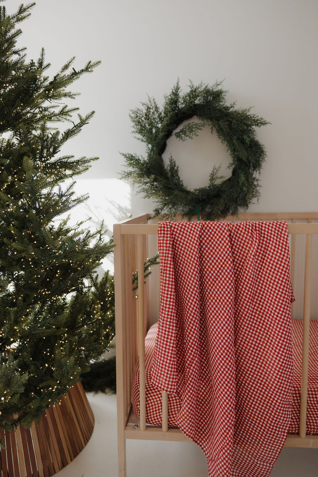 A wooden crib with a forever french baby Muslin Swaddle in Red Gingham is draped over it beside a lit Christmas tree. A green wreath hangs on the white wall above the crib.