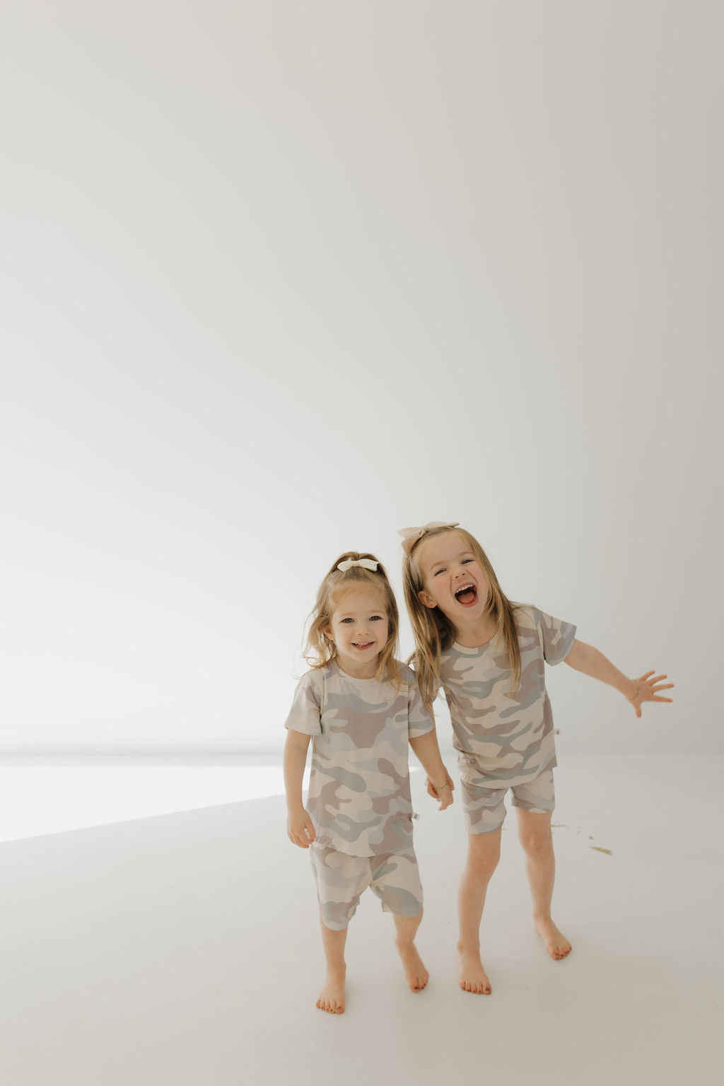 Two young kids in matching "Bamboo Short Set | Camo" pajamas by forever french baby stand barefoot on a light floor. One laughs with an extended arm, and the other smiles gently against a bright plain wall, showcasing their comfy and stylish breathable sleepwear.