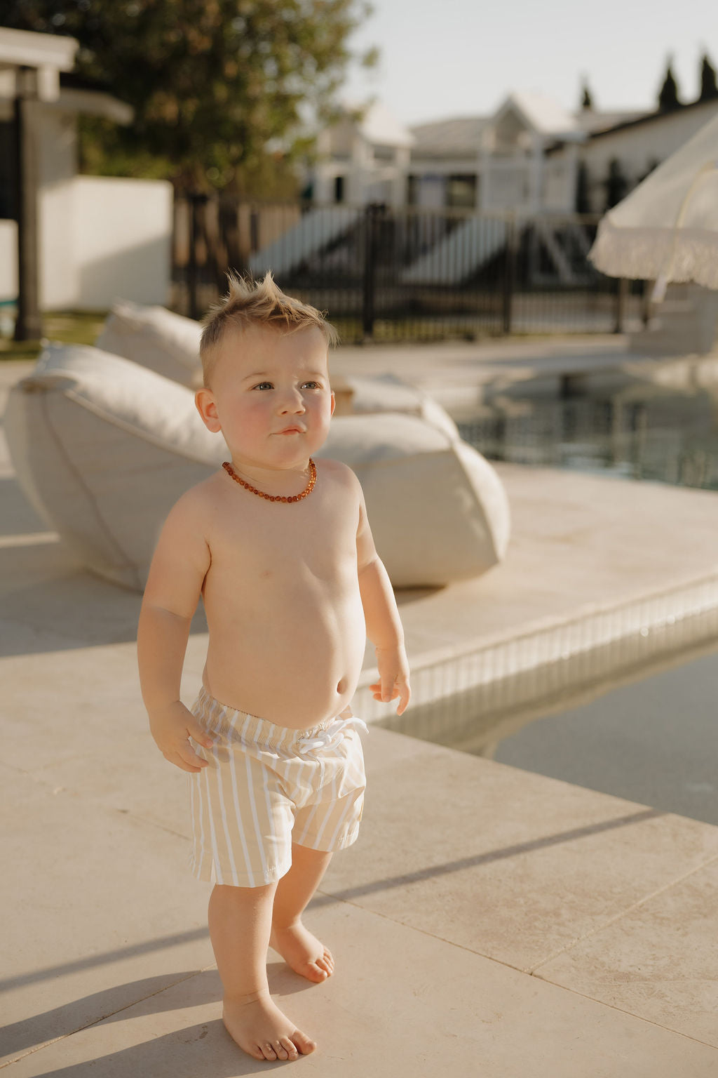 A young child, barefoot by a pool with outdoor furniture and modern homes behind, wears lolo webb 2's Child Board Short | Sun Rays and an amber necklace.