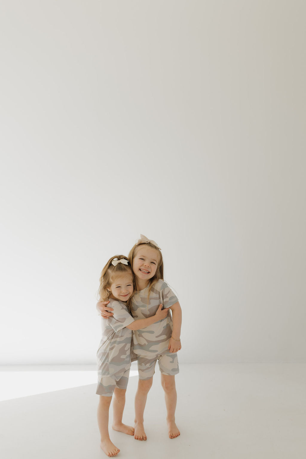 Two young children, barefoot on a light floor, wear matching Bamboo Short Sets in Camo from forever french baby. The older child lovingly hugs the younger, both smiling joyfully against a plain, softly lit wall.