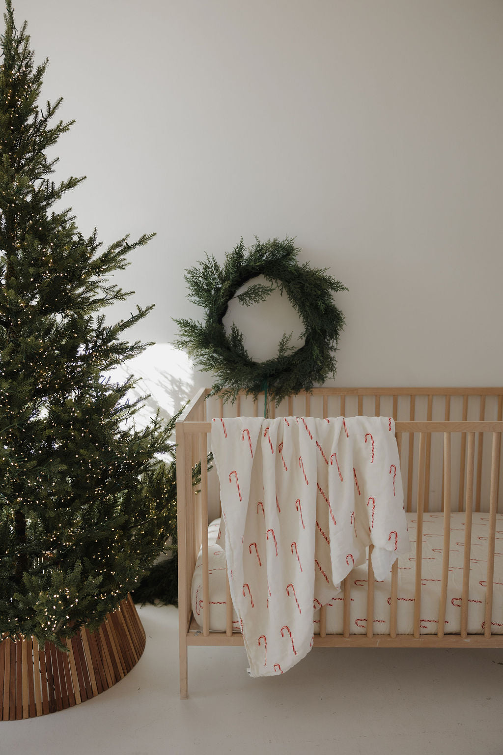A minimalist nursery with a wooden crib, a forever french baby Muslin Quilt | Candy Cane draped over the side, a Christmas wreath on the wall, and a lit Christmas tree beside the crib.