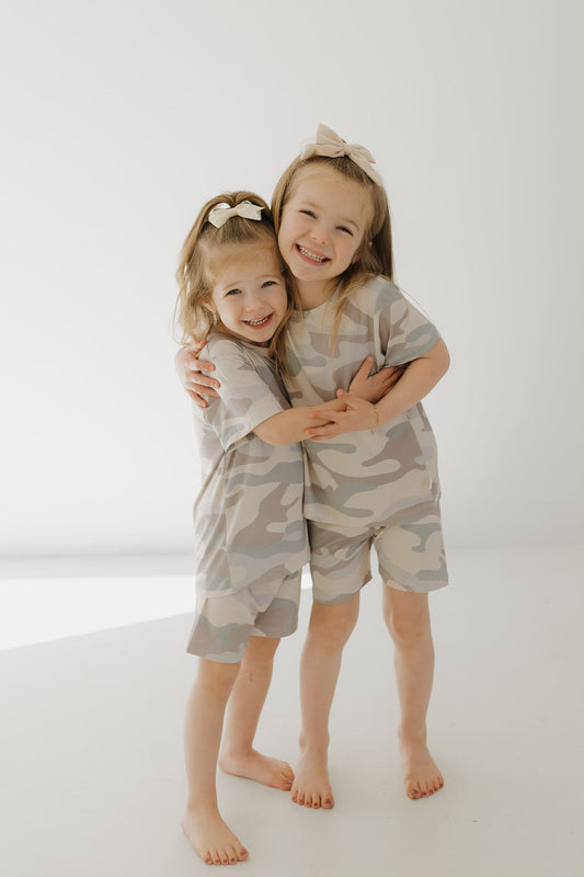 Two young children smile and hug in matching neutral-toned Bamboo Short Sets | Camo by forever french baby. Their hypo-allergenic clothing ensures comfort, while bows adorn their hair as they stand barefoot against a plain, light-colored backdrop.