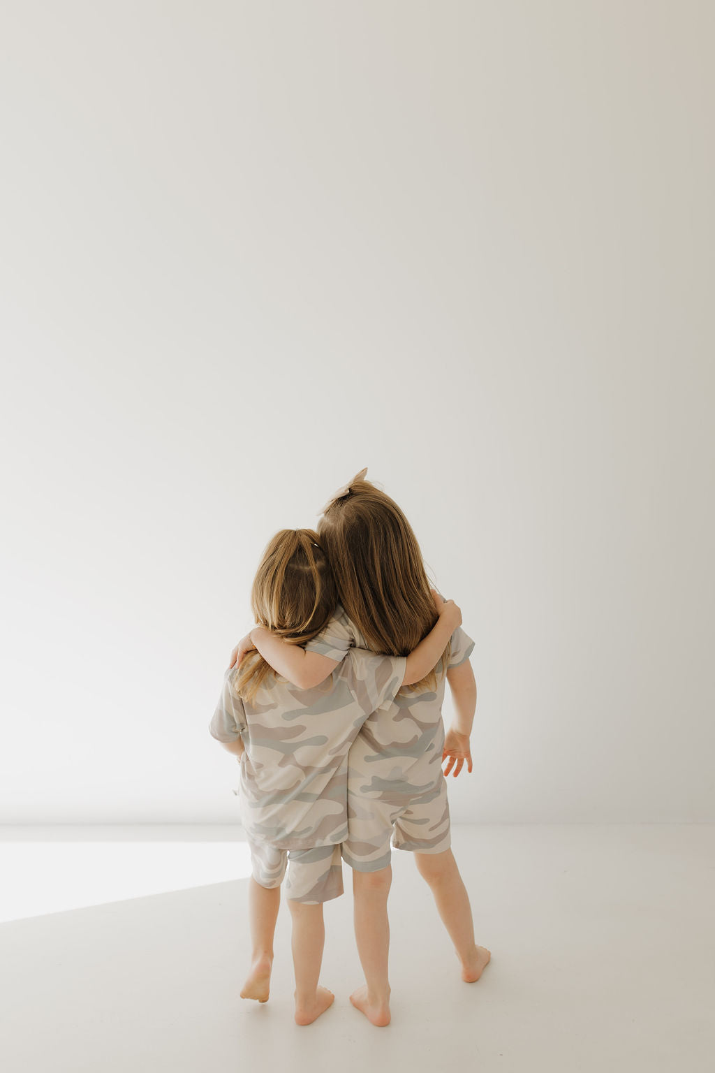 Two children with long hair, in matching "Bamboo Short Set | Camo" outfits from forever french baby, stand and embrace each other. They face away from the camera against a plain white background.