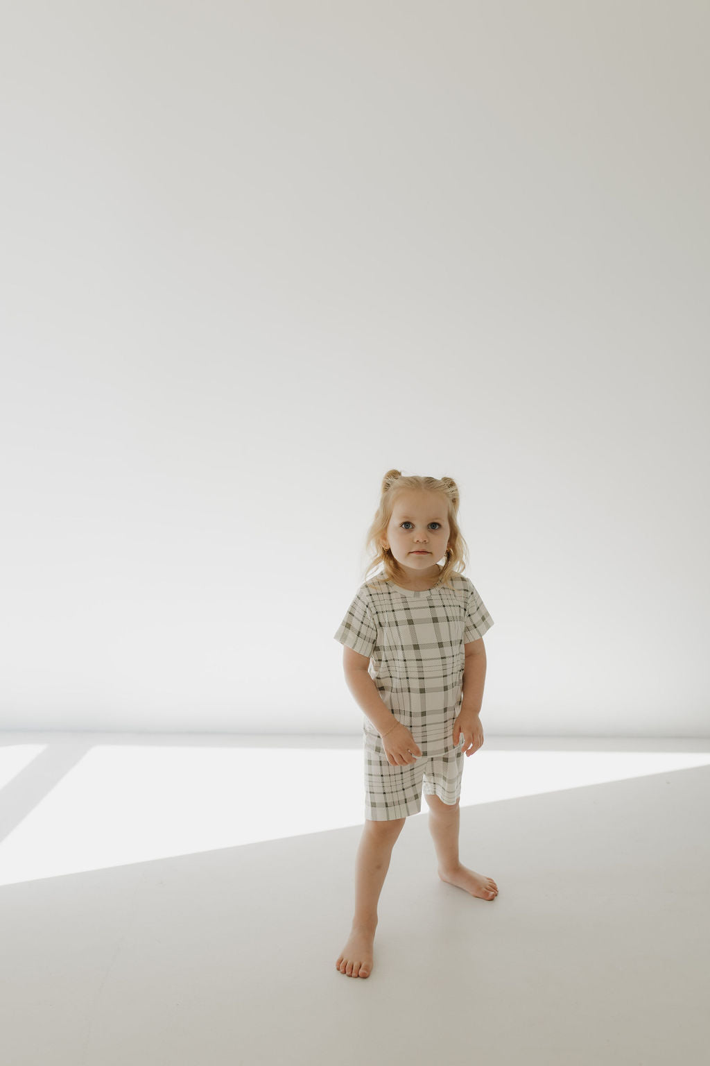 In a bright, minimalistic room, a young child with light hair stands barefoot, wearing the Bamboo Short Set in Picnic Plaid by forever french baby. The breathable outfit complements the serene setting as soft sunlight and shadows play across the plain background.