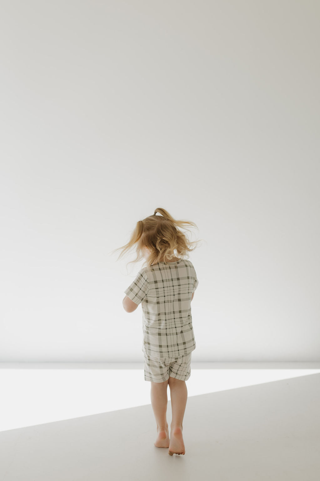A young child with light brown hair, dressed in the Forever French Baby Bamboo Short Set in Picnic Plaid, stands barefoot on a white floor. The bright and minimalist background creates a serene and airy atmosphere.