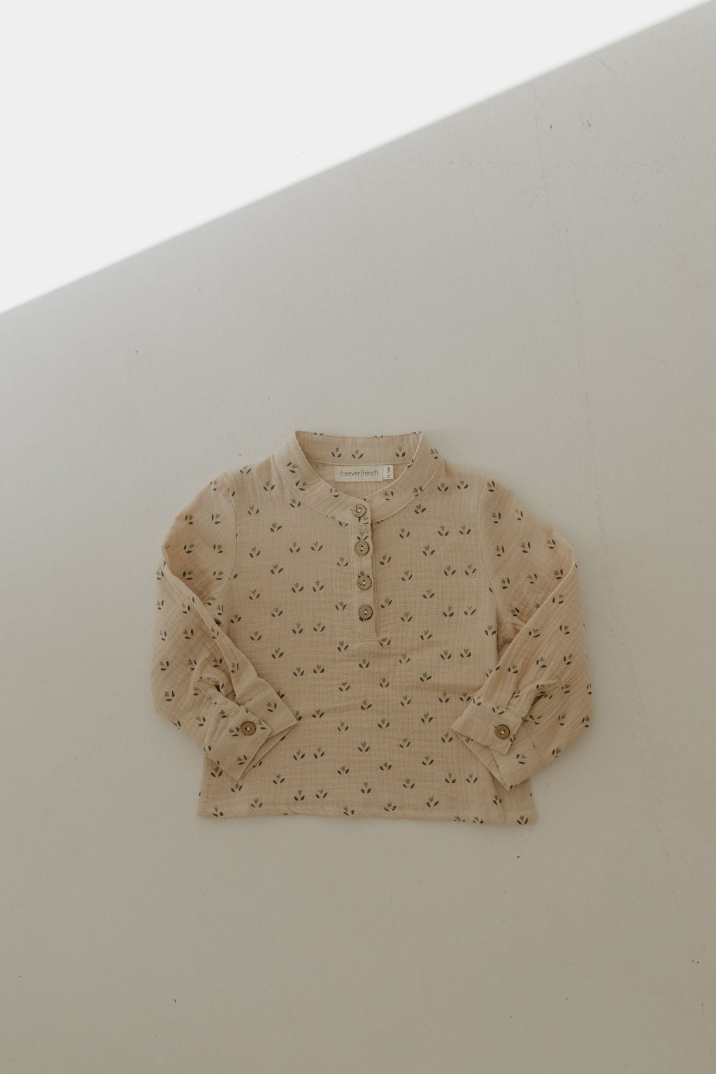 The Muslin Button Up | Flora by forever french baby is a beige, long-sleeve muslin cotton shirt with a floral pattern, buttoned cuffs, and partial placket, laid flat in sunlight. Perfect for kids' collections.