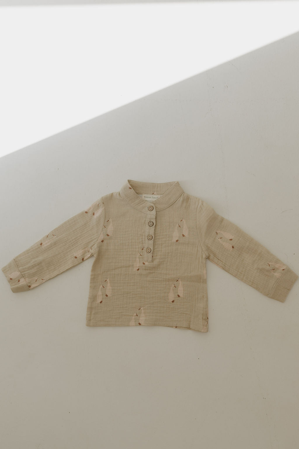 The forever french baby Muslin Button Up | Silly Goose is a beige long-sleeve 100% cotton muslin shirt with a mandarin collar, four-button front, and subtle animal print, shown flat on a light surface with sunlight across the top.