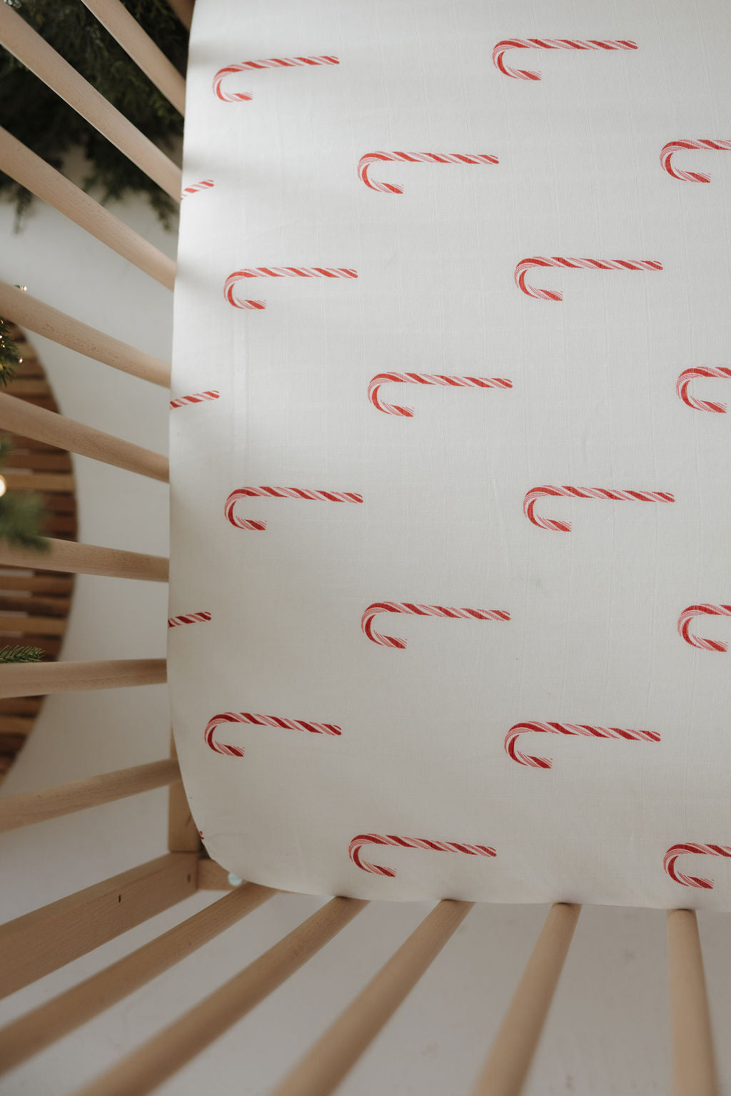 The Muslin Crib Sheet | Candy Cane (Preview) by forever french baby features a festive red and white candy cane pattern, shown covering a crib mattress with wooden slats visible—ideal for holiday-inspired nursery bedding.