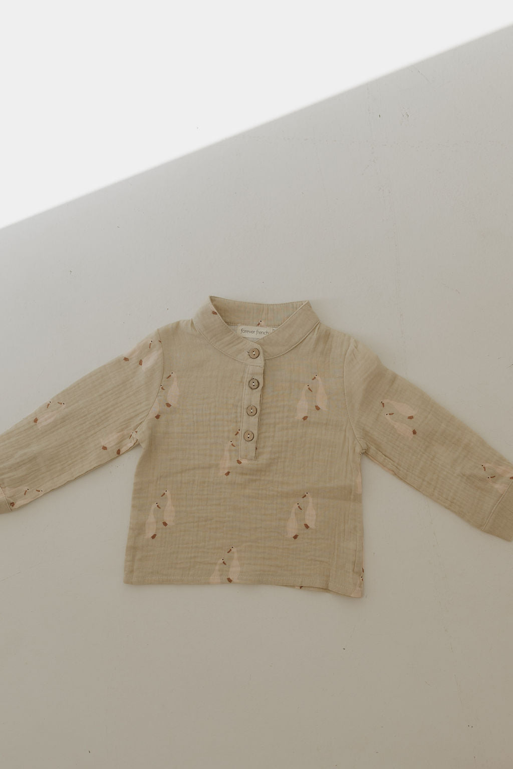 The Muslin Button Up | Silly Goose by forever french baby is a beige, long-sleeve baby shirt made from 100% cotton muslin with a mandarin collar, front buttons, and an animal face pattern—soft, breathable style. Laid flat on white.