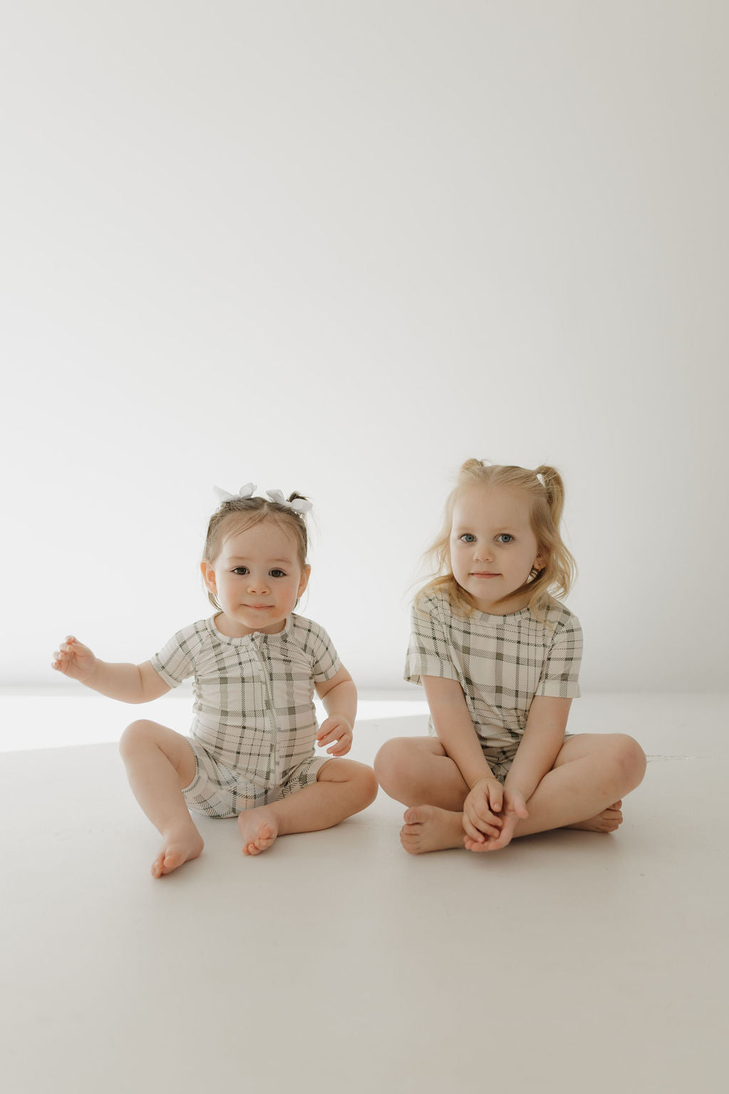 Two young children sit on a light-colored floor against a plain background, wearing matching outfits from forever french baby's Bamboo Short Set in Picnic Plaid. The child on the left sits with legs outstretched, while the one on the right sits cross-legged, both looking toward the camera.