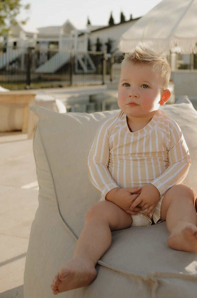 A young child with short blond hair sits on a beige outdoor cushion, wearing the lolo webb 2 Toddler Rash Guard | Sun Rays. The background features a pool area with white structures and greenery in soft sunlight.