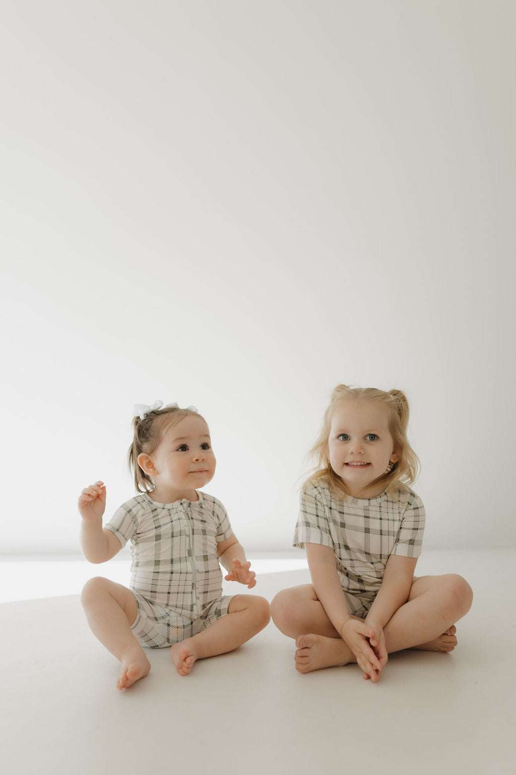 Two young children sit on a white floor with a plain backdrop, dressed in matching "Bamboo Short Set | Picnic Plaid" by forever french baby. The child with shorter hair has hands on the floor, while the other has slightly longer hair, clasps hands together, and smiles.