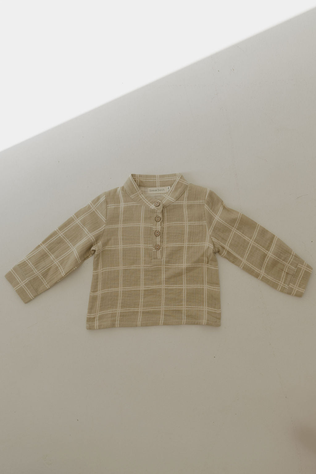 The Muslin Button Up | Linen Grid by forever french baby is a beige, collarless long-sleeve shirt with a white plaid pattern and three front buttons, made from soft muslin cotton. Shown laid flat on a light surface with a diagonal shadow across the top.