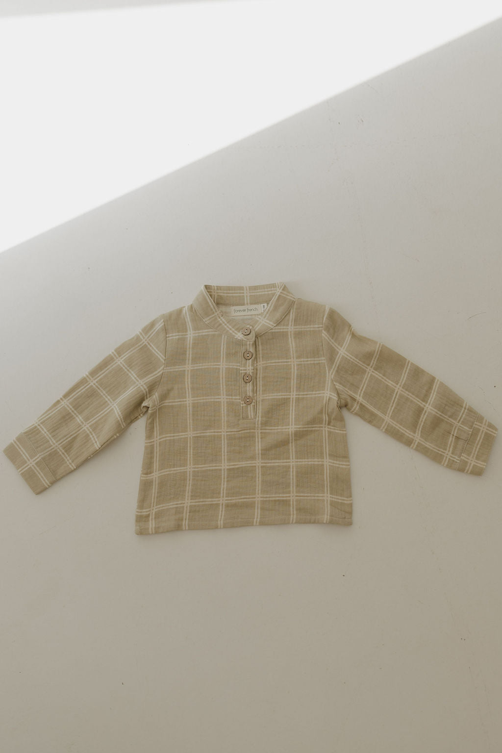 The forever french baby Muslin Button Up | Linen Grid is a beige and white plaid long-sleeve toddler shirt made from breathable muslin cotton, featuring a collar and three front buttons, shown laid flat on a light-colored surface.