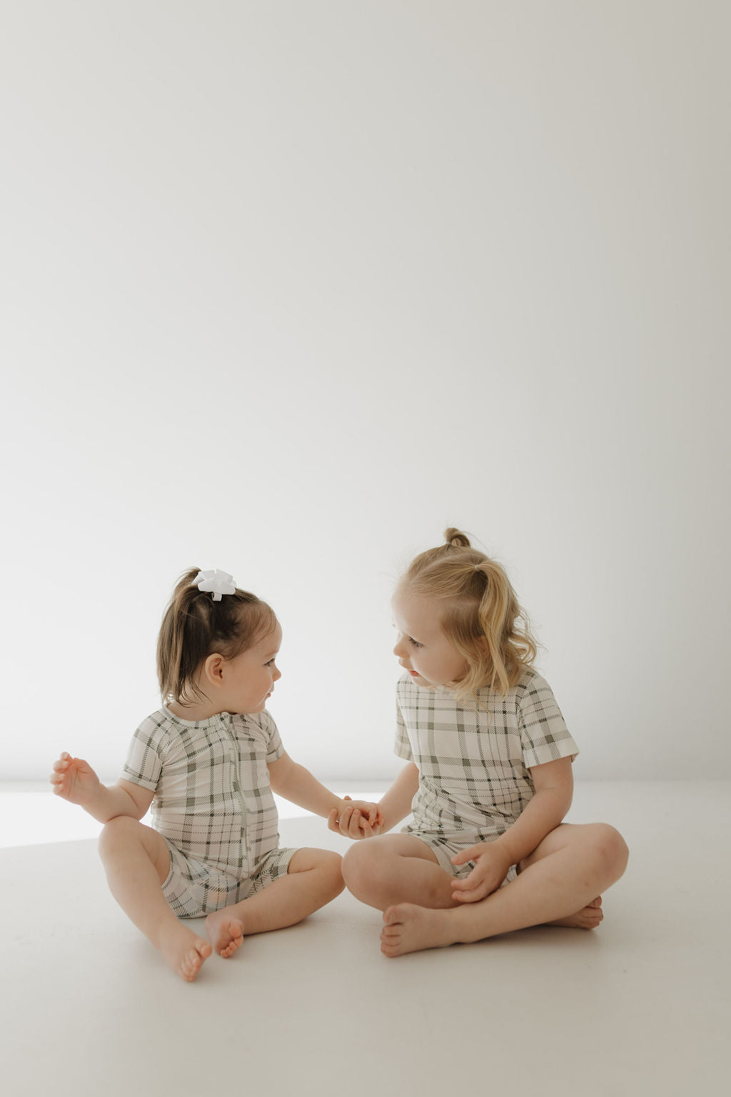 Two young children sit on the floor in front of a light backdrop, wearing matching forever french baby Bamboo Short Set | Picnic Plaid outfits. The child on the right has a ponytail, while the left sports a white bow. They hold hands and gaze at each other playfully.