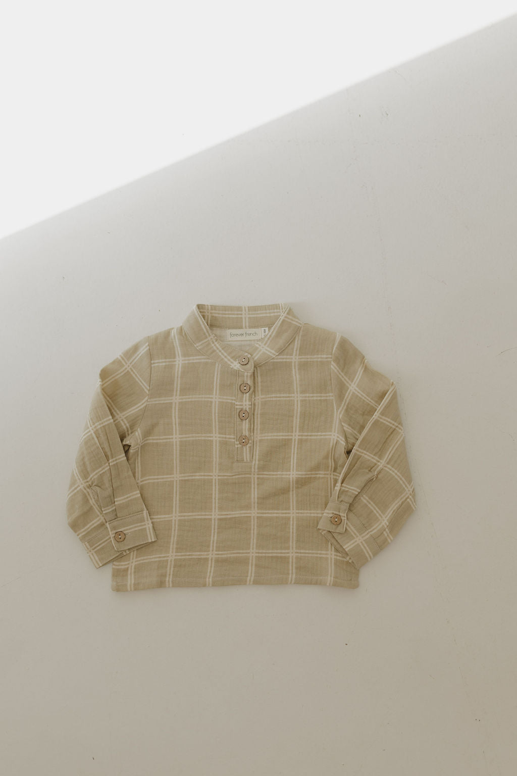 The Muslin Button Up | Linen Grid by forever french baby, a beige plaid long-sleeve shirt in breathable muslin cotton with buttoned neck and cuffs, is laid flat on a light surface beneath gentle, diagonal sunlight.