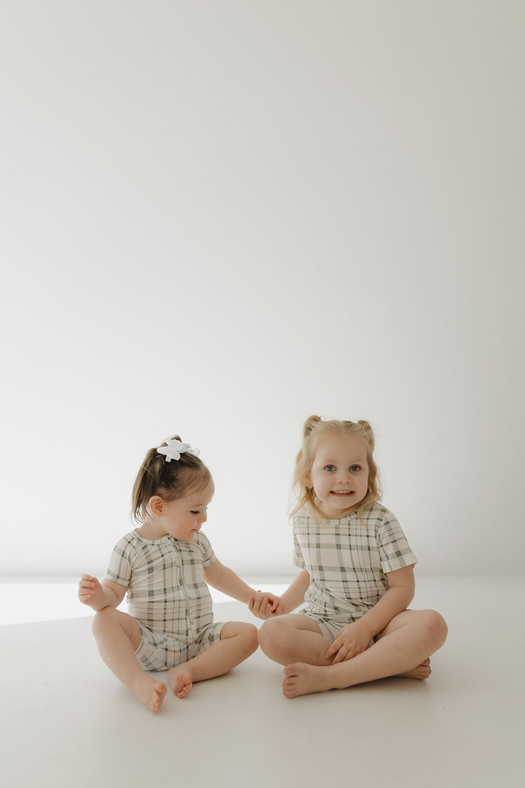 Two young children sit on the floor against a plain white background, wearing matching forever french baby Bamboo Short Sets in Picnic Plaid. One child smiles at the camera while the other gazes downward. Both have light hair, and the smaller child sports a bow.