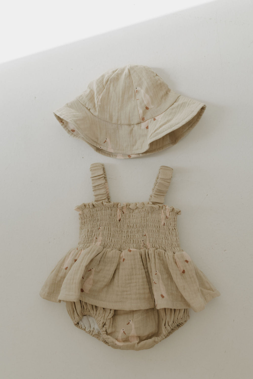 The Sleeveless Muslin Romper & Hat by forever french baby is a beige 100% cotton romper set with ruched straps, ruffle details, a bucket hat, and pink floral print, shown laid flat on a white background.