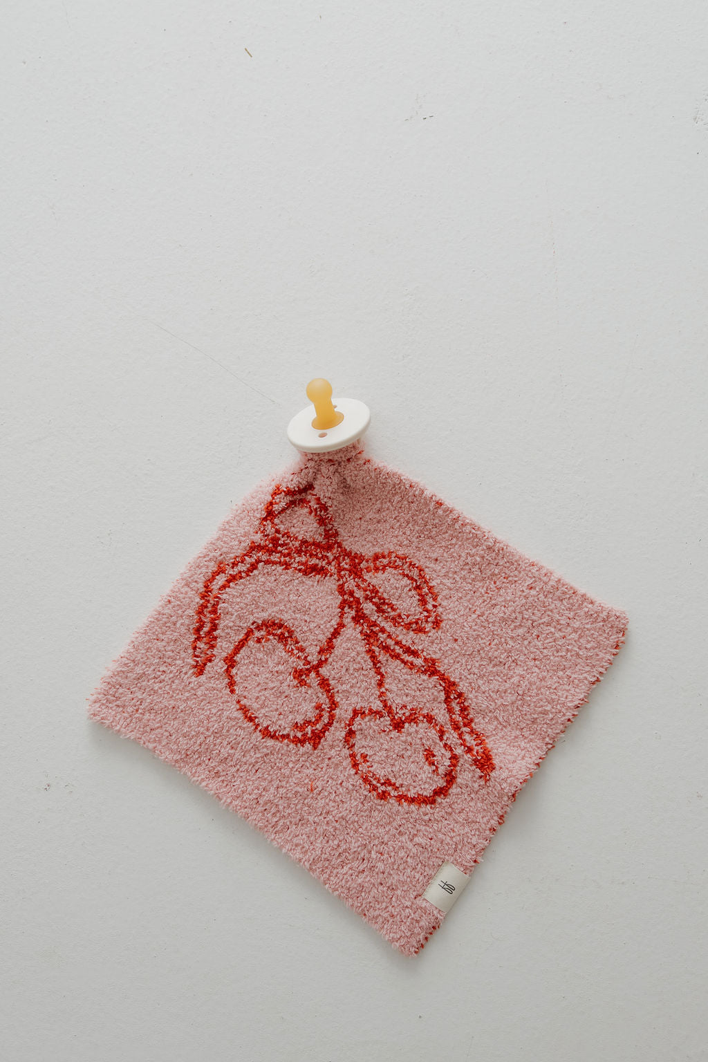 The Minty Cherries Lovey | Iconic Cherry, a pink textured plush with red cherry print and an attached yellow-white pacifier, rests on a light gray surface.