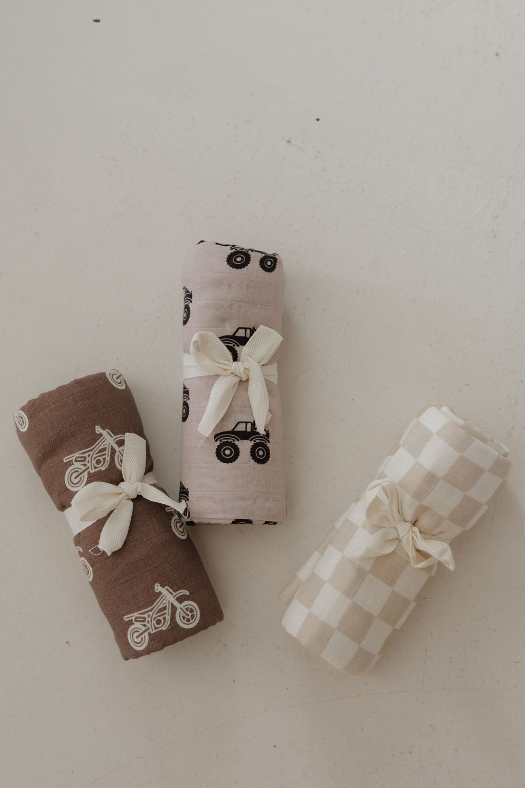 Three rolled forever french baby blankets, including the Muslin Swaddle | Checkerboard and two other printed swaddles, are tied with ribbon and displayed on a light surface.