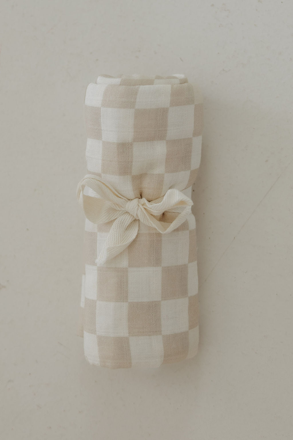 The Muslin Swaddle | Checkerboard by forever french baby, featuring a beige and white gingham pattern, is rolled up and tied with a cream ribbon, displayed on a light, textured surface.