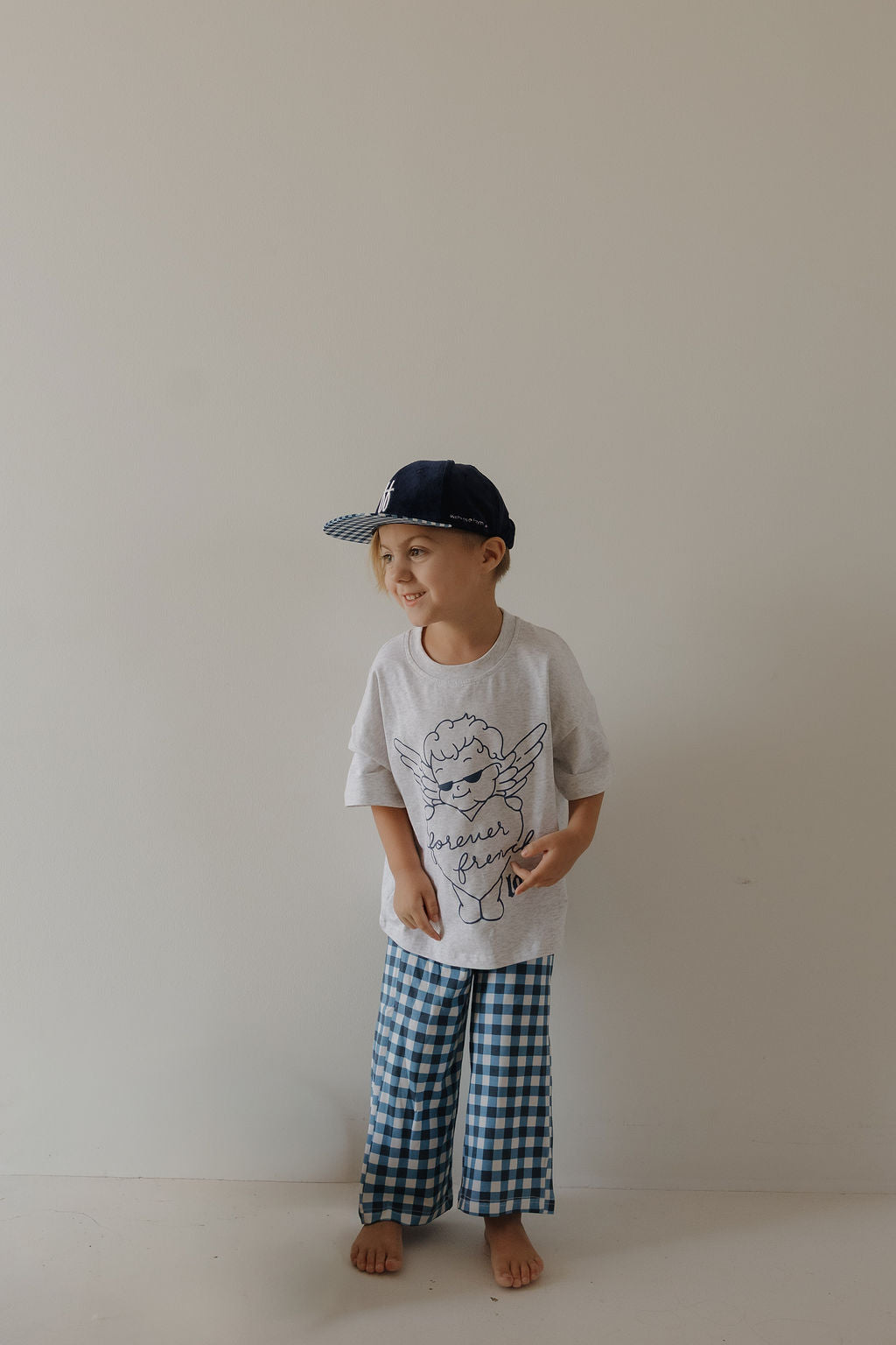 A young child wearing the forever french baby Snapback | Blue Corduroy ff stands barefoot against a plain light-colored wall, smiling and looking to the side.