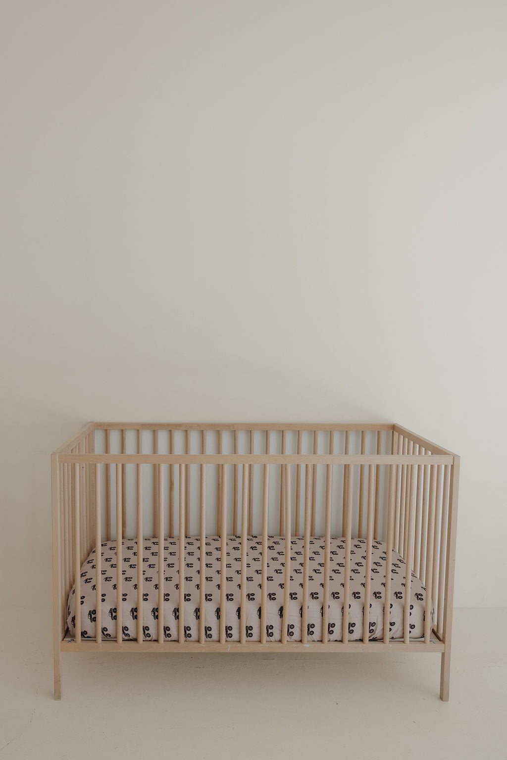 A minimalist wooden baby crib with a forever french baby Muslin Crib Sheet in the Monster Truck print, set against a plain off-white wall.