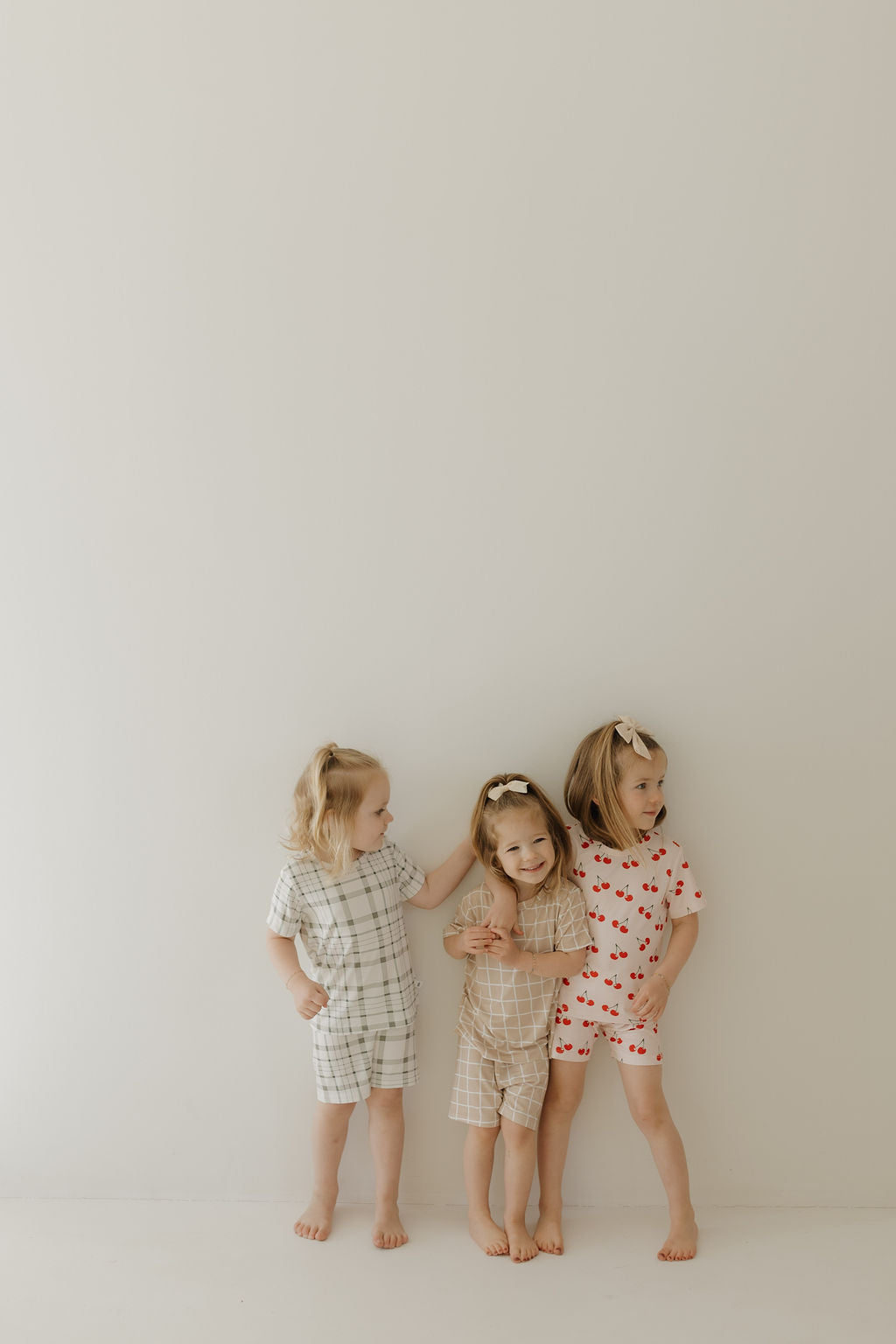 Three young children in matching outfits stand against a plain, light-colored wall. Two wear the Bamboo Short Set | Grid by forever french baby, and one wears a strawberry-patterned outfit. Their breathable clothing and playful interactions create a joyful scene as they smile together.