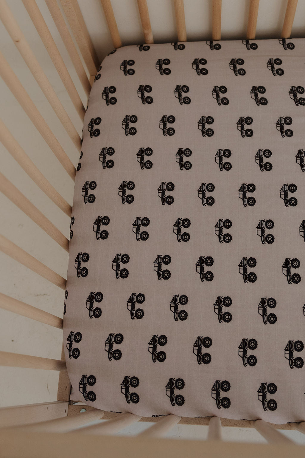 A close-up of a crib with light wooden slats featuring the forever french baby Muslin Crib Sheet | Monster Truck, decorated with a black and white monster truck pattern on a beige background.