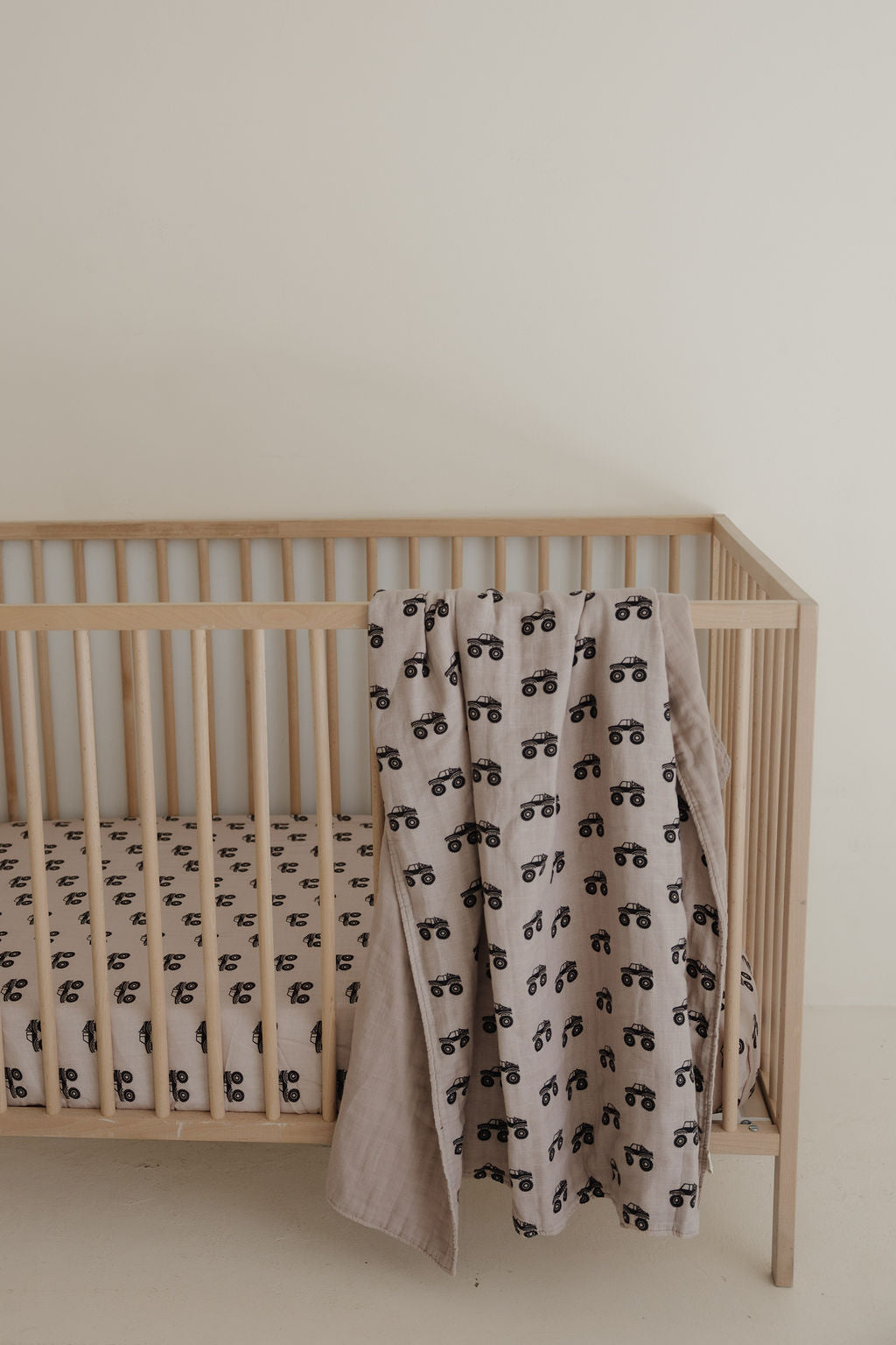 A light wooden crib with a forever french baby Muslin Crib Sheet featuring a monster truck print, paired with a matching blanket, set against a plain cream-colored wall.