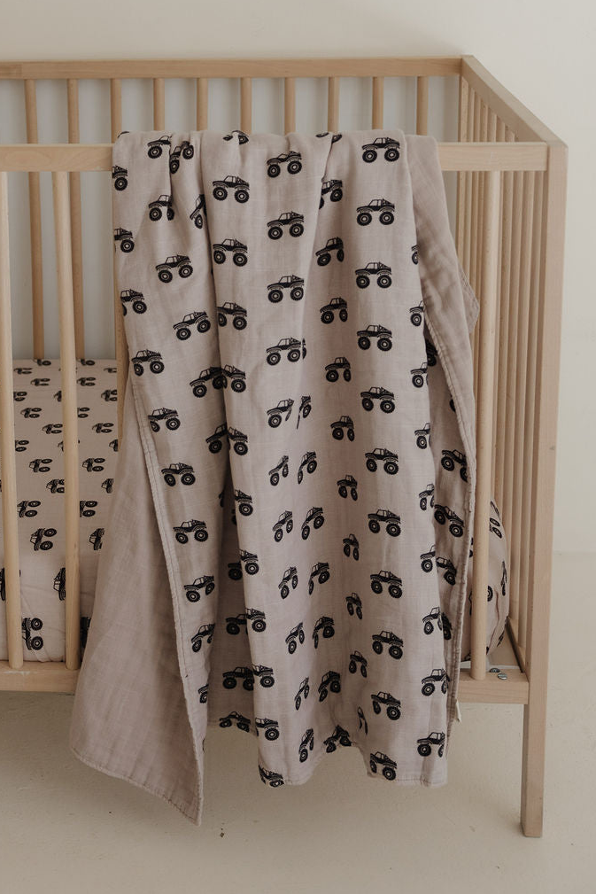 A light brown Muslin Quilt | Monster Truck by forever french baby, featuring a black monster truck pattern, hangs over a wooden crib with matching nursery bedding in a minimal, neutral setting.