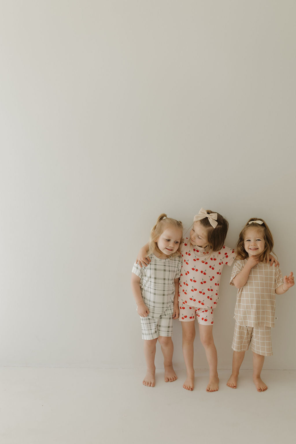 Three young children stand against a light background, smiling. They wear forever french baby Bamboo Short Sets with grid, cherry, and neutral patterns made from hypoallergenic fabric. Two have bows in their hair.